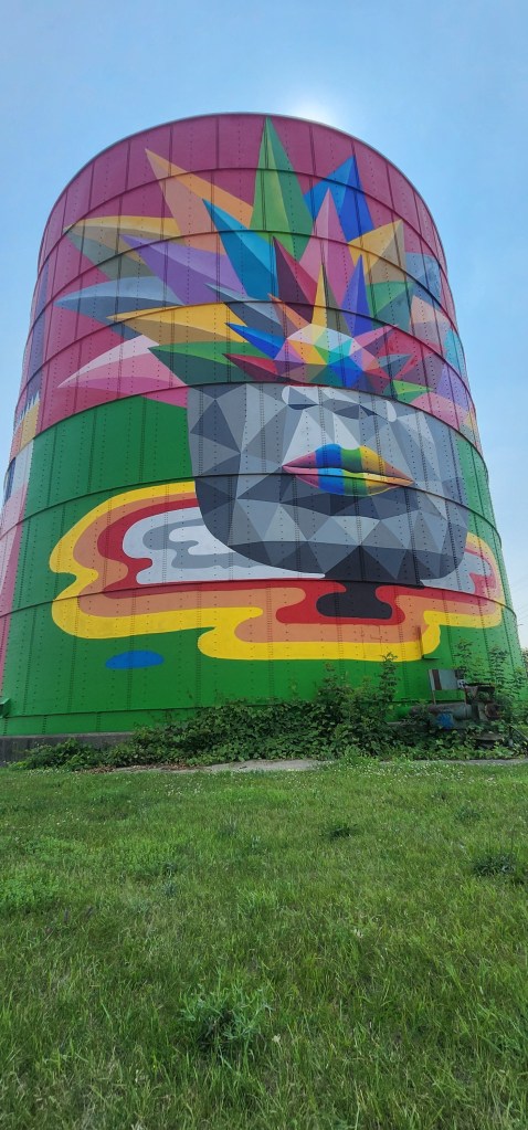 A brightly colored mural on a large grain silo featuring geometric shapes, vibrant patterns, and a stylized face with a colorful explosion of shapes above it, set against a clear blue sky.