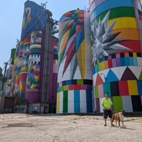 A colorful mural on old grain silos with vibrant geometric patterns, featuring a person in a yellow shirt walking a dog in front of them.
