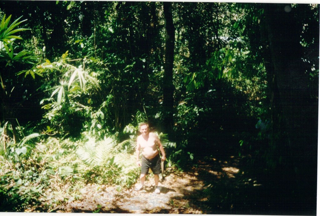 A person stands in a dense rainforest, surrounded by lush green plants and trees, wearing shorts and holding a walking stick.