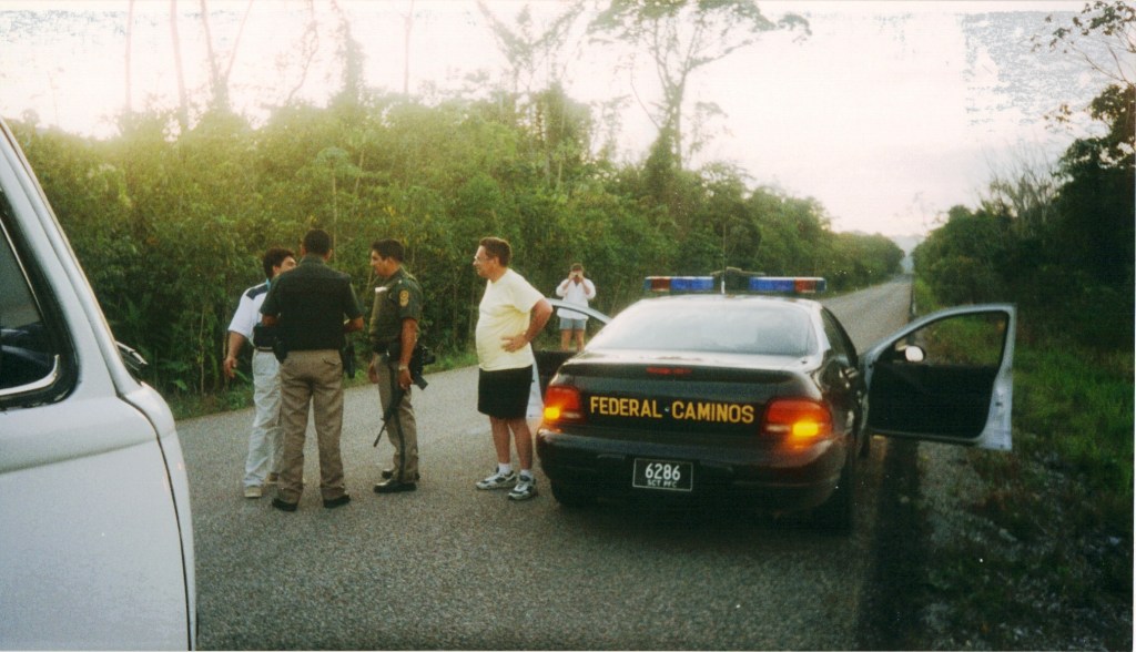 Law enforcement officers conversing on a rural road in Chiapas, Mexico, with a police car marked 'FEDERAL CAMINOS' parked nearby.