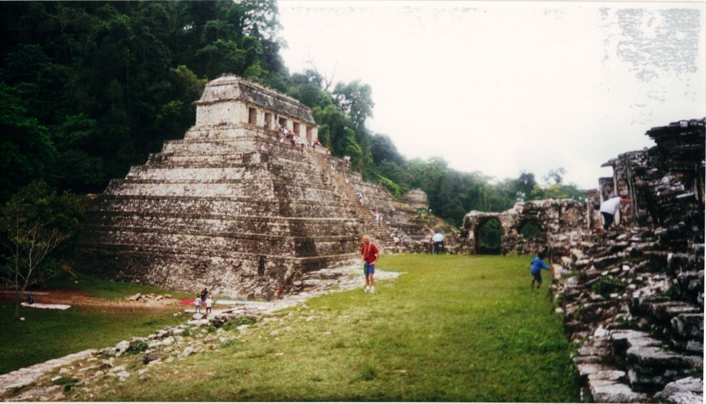 A glimpse of the ancient ruins at Palenque, featuring a stepped pyramid surrounded by lush greenery and visitors exploring the site.