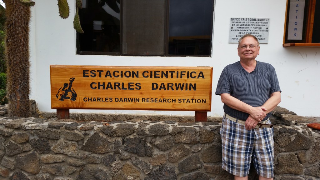 A person standing in front of the Charles Darwin Research Station sign, which reads 'Estacion Cientifica Charles Darwin', with a stone wall in the foreground.
