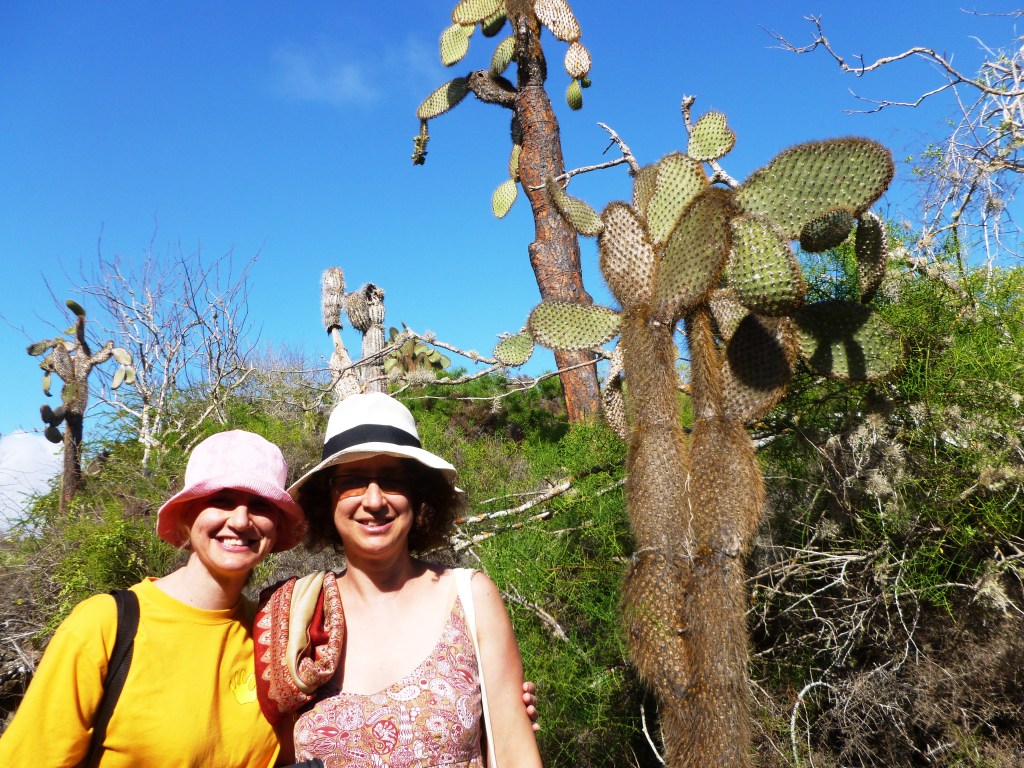 Two women stand next to a giant cactus in the Galapagos Islands, surrounded by lush greenery and a clear blue sky.