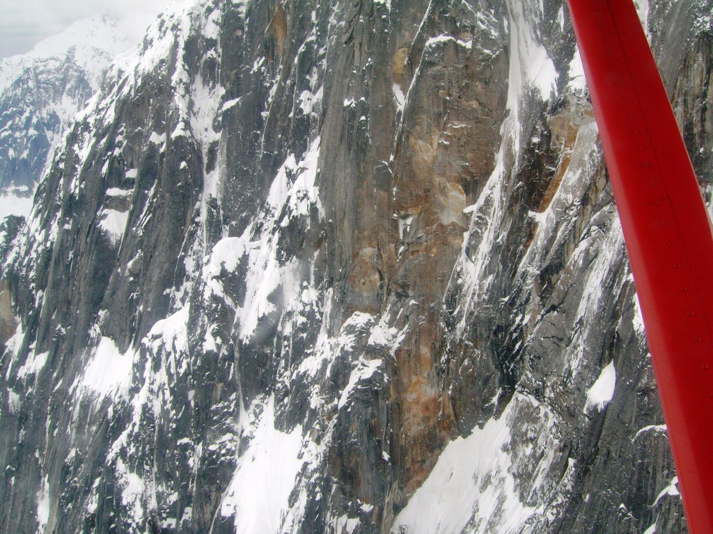 Aerial view of rocky mountain cliffs covered in snow with a red airplane wing in the foreground.