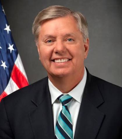 Portrait of a smiling man in a black suit and a blue and white striped tie, standing in front of an American flag.