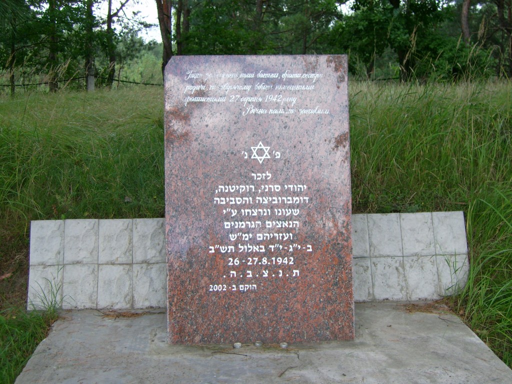 A granite memorial stone featuring Hebrew and Russian inscriptions, commemorating Jewish victims from a specific historical event. Surrounded by greenery in a natural setting.