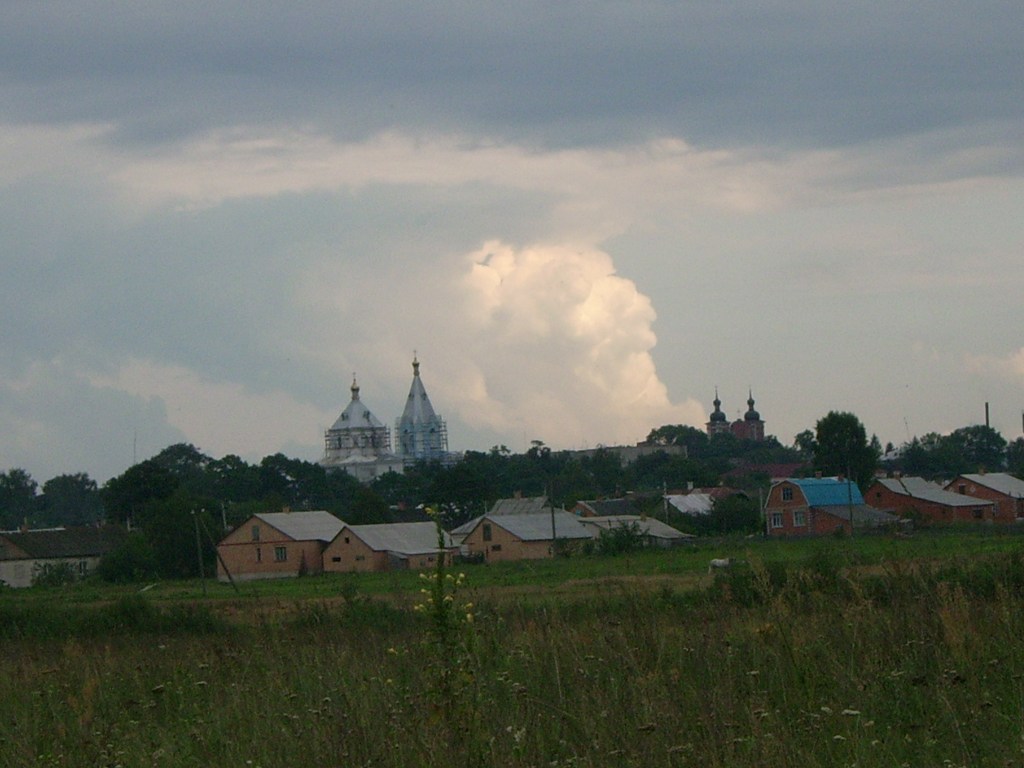 A rural landscape featuring clouds overhead, with two distinct religious buildings in the background, surrounded by scattered houses and greenery.
